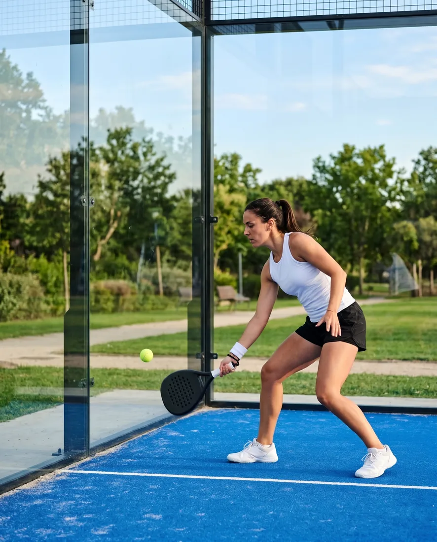 A woman plays the ball as it comes back off the clear glass wall of a park court