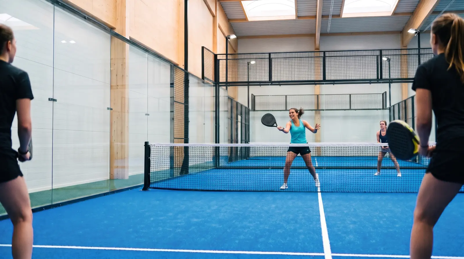 A smiling woman hits a volley in a bright indoor padel hall